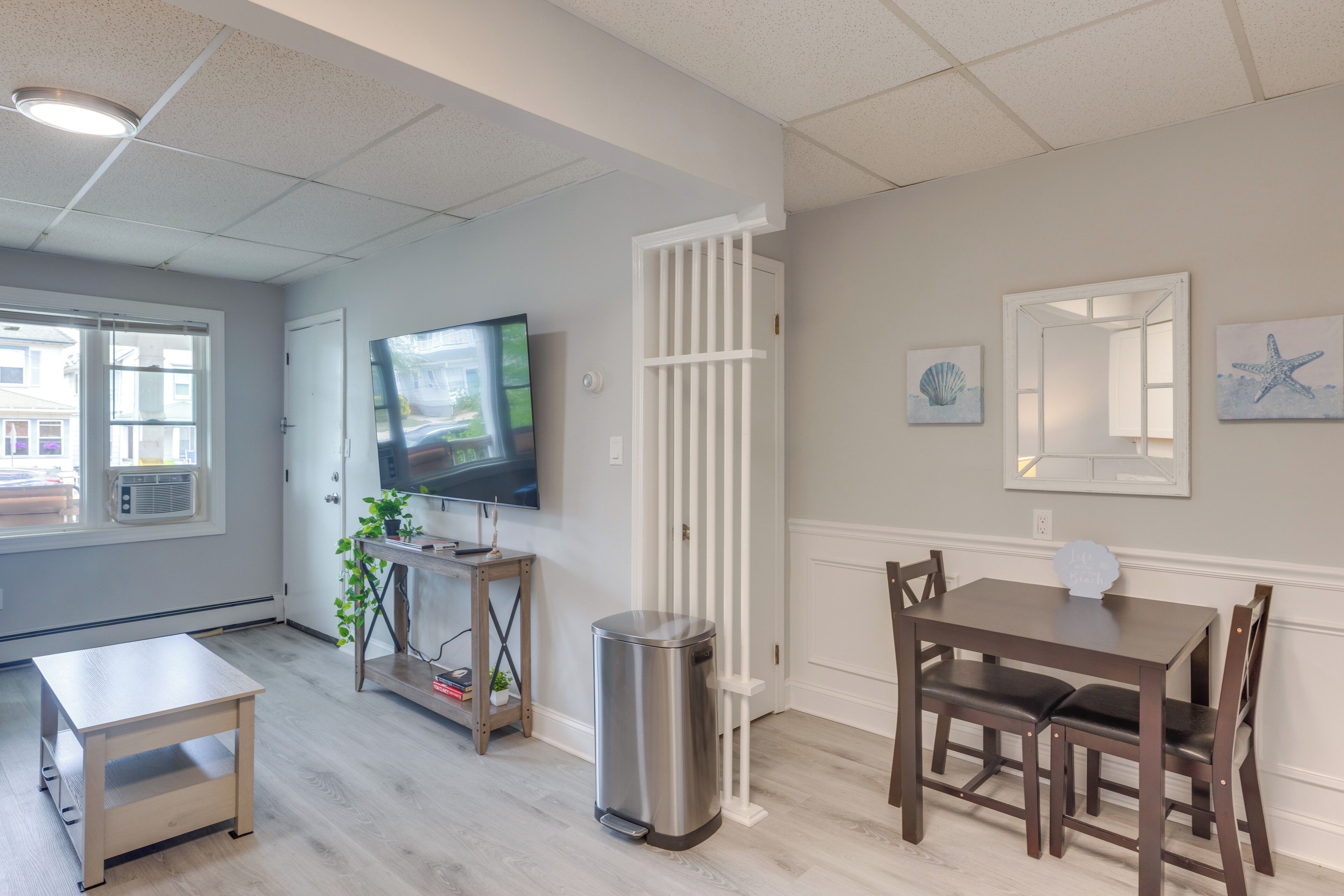 A modern living room with a flat-screen TV, a dining table with chairs, a trash can, and a window with blinds.