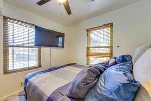 Une chambre à coucher comprenant un lit, un ventilateur de plafond, un téléviseur à écran plat fixé au mur et deux fenêtres équipées de stores.