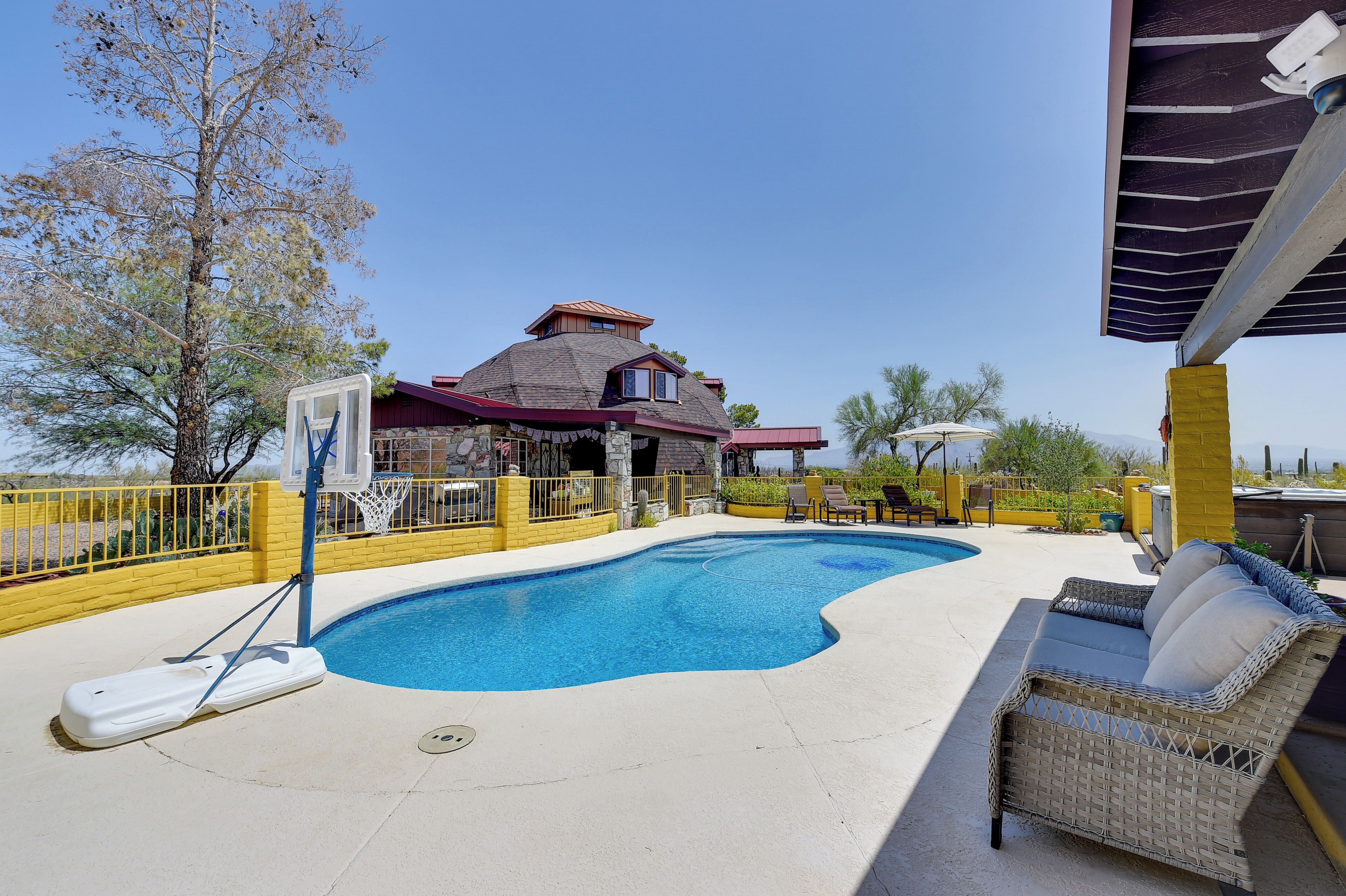 A swimming pool with a basketball hoop, a covered patio area with chairs, and a house with a red roof.