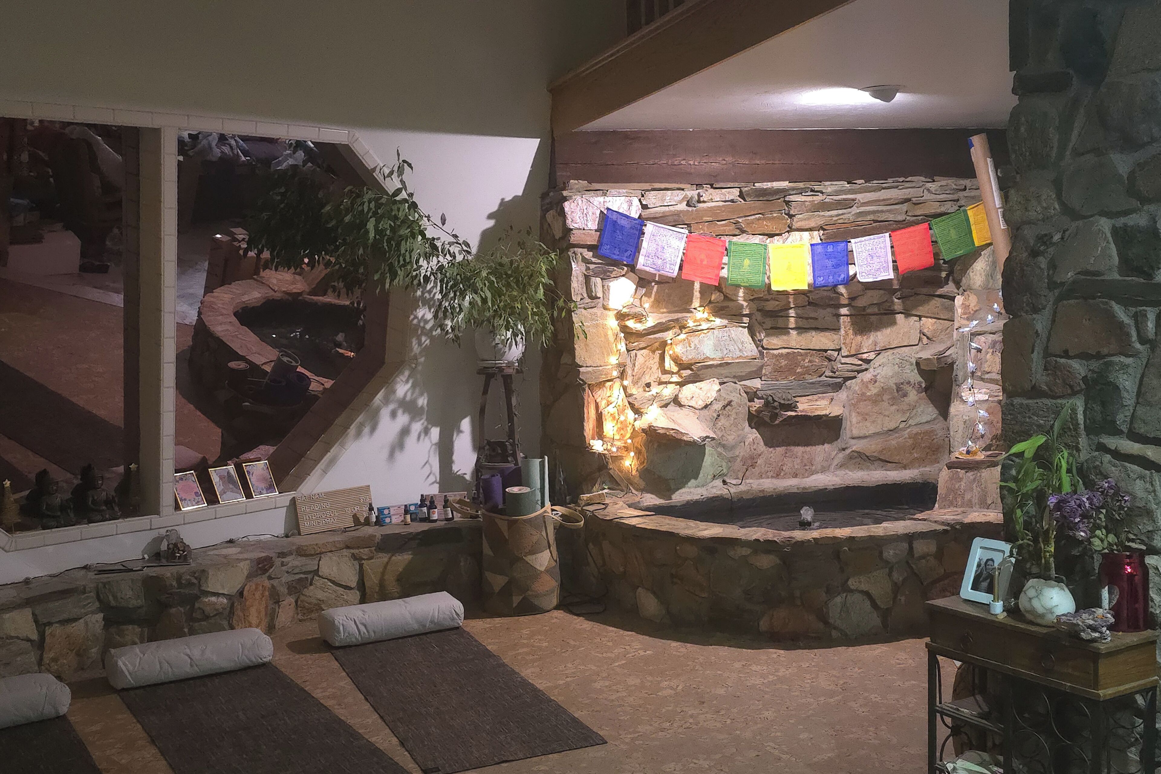 A stone fireplace with colorful prayer flags, a stone bench, and a small table with a vase and picture frame.