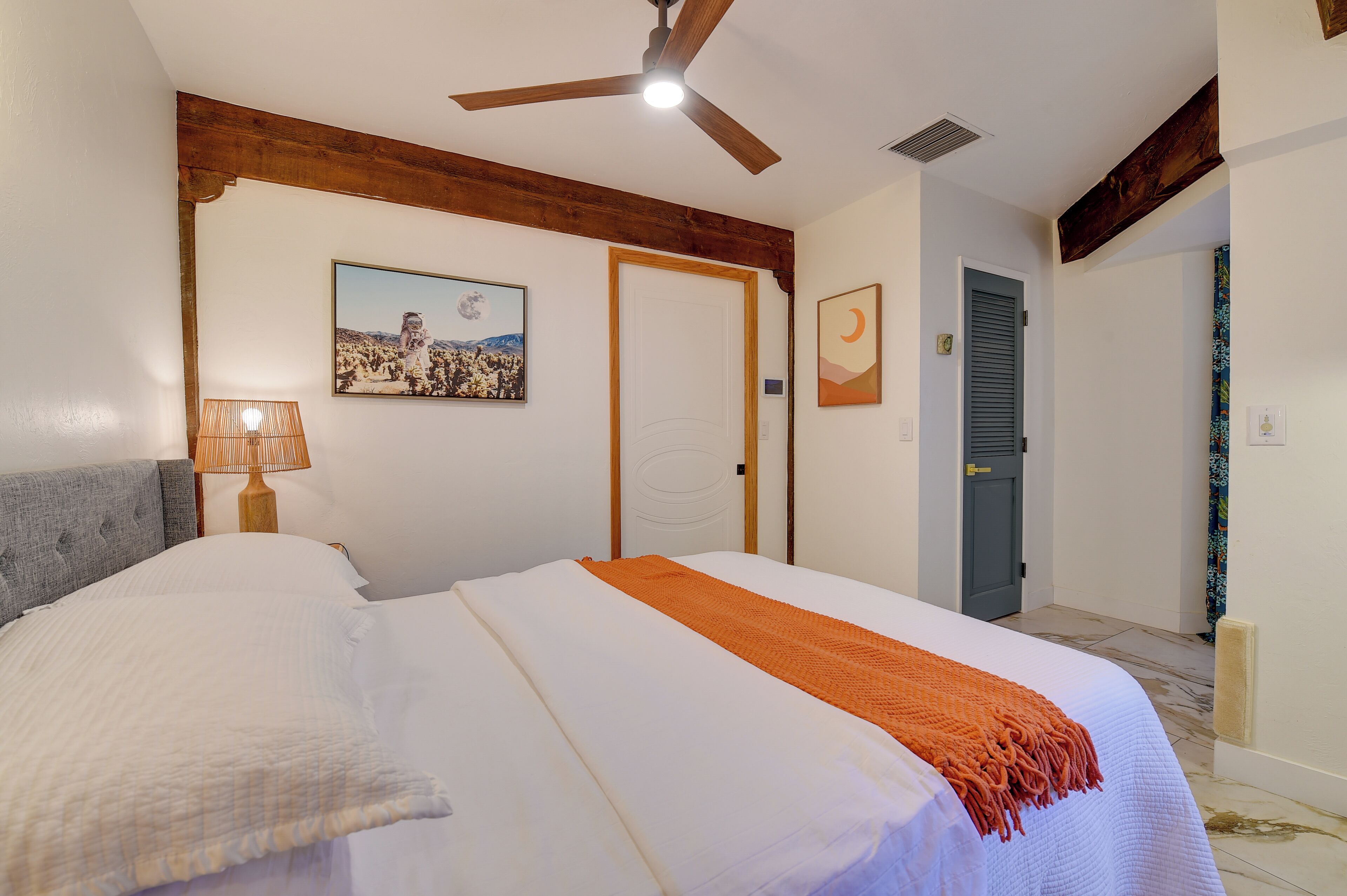 A double bed with a white and orange blanket, a wooden headboard, a ceiling fan, and a wall-mounted lamp.