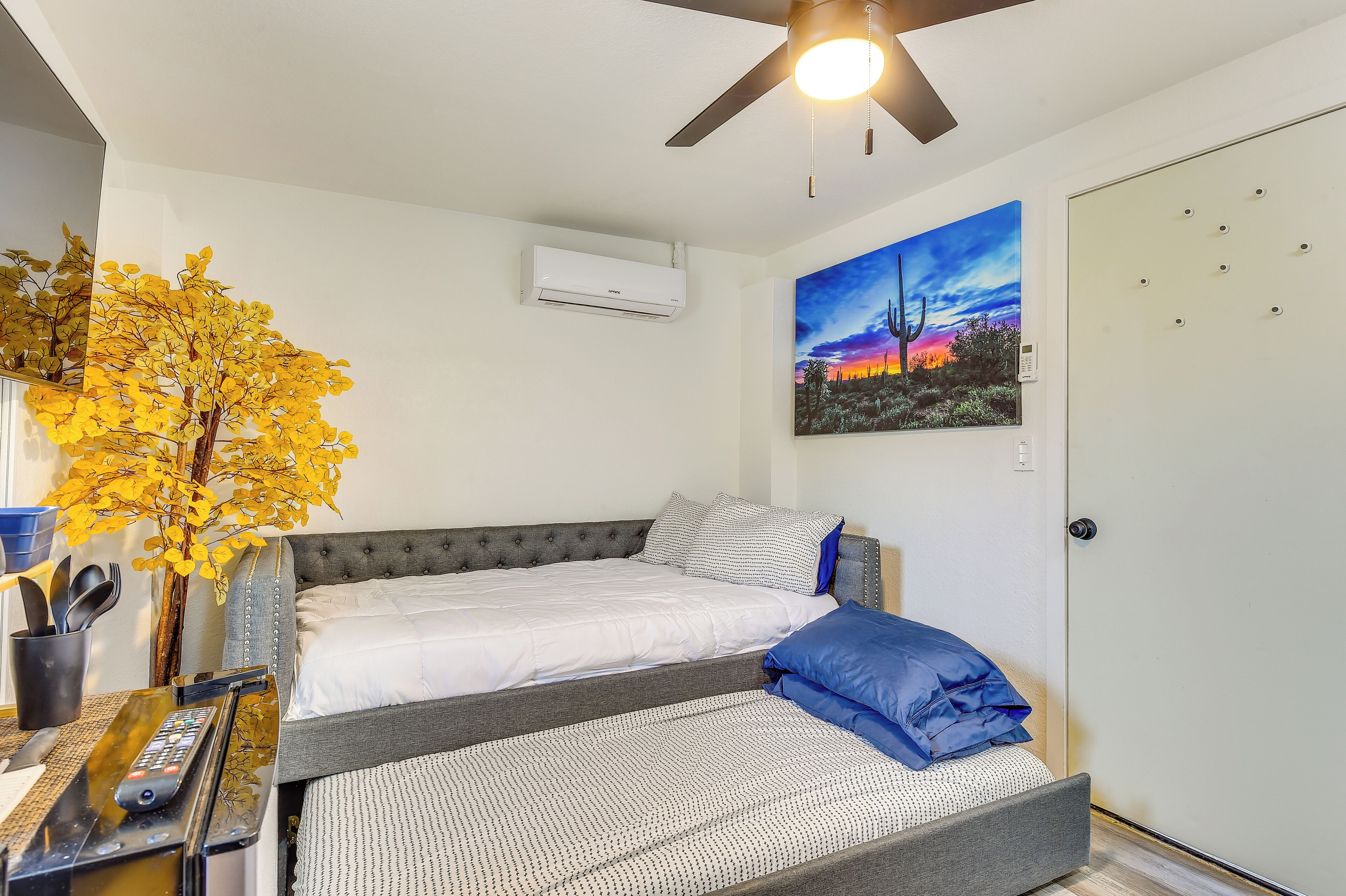 Un dormitorio con cama, ventilador de techo, aire acondicionado, un cuadro y una planta en maceta.