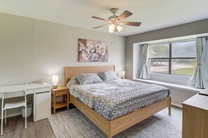 Ein Schlafzimmer mit Bett, Schreibtisch, Stuhl, Deckenventilator, einem Fenster mit Vorhängen und einem Bild an der Wand.