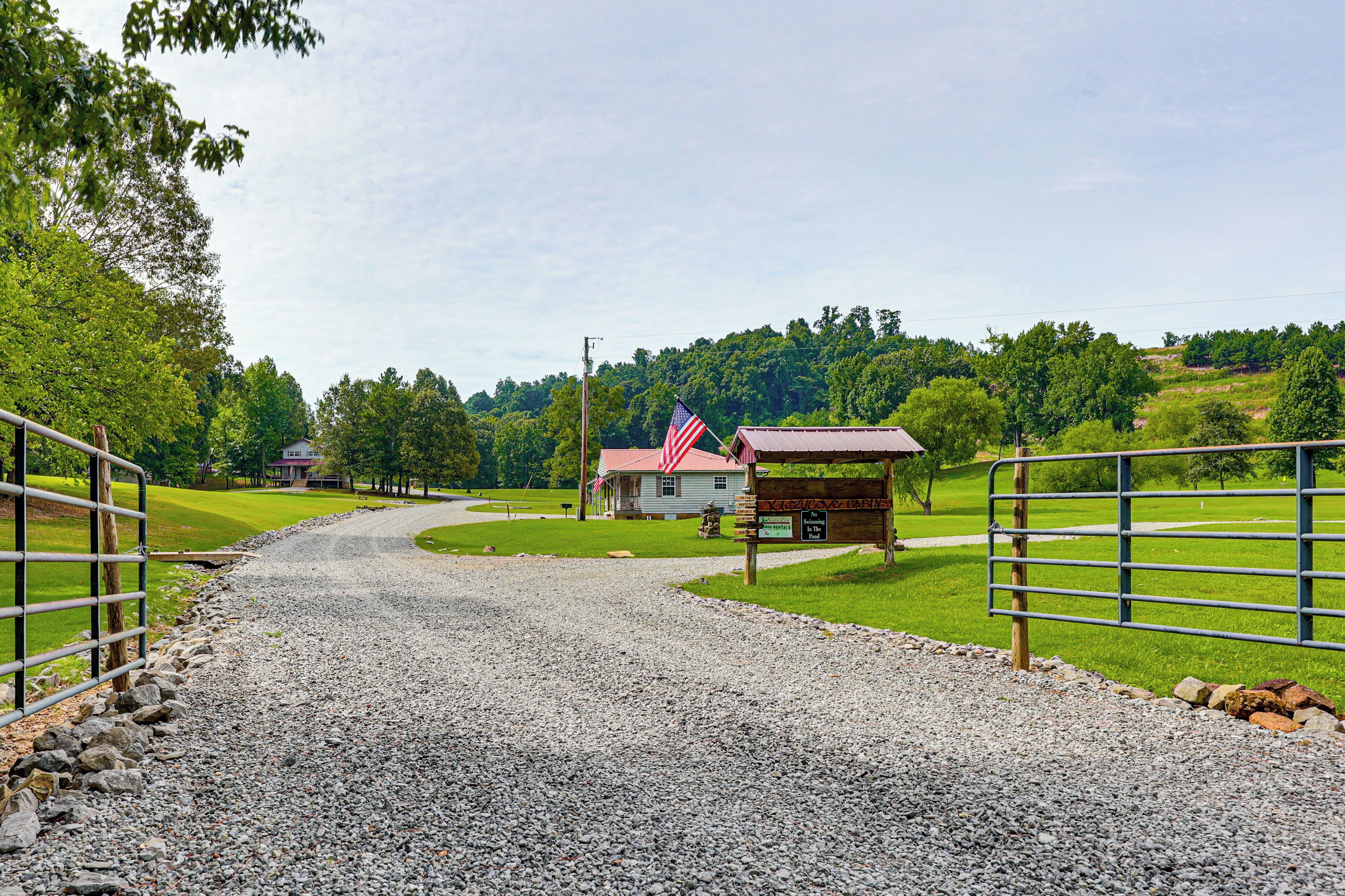 A gravel driveway leading to a rustic cabin with an American flag, surrounded by lush greenery and a clear sky.
