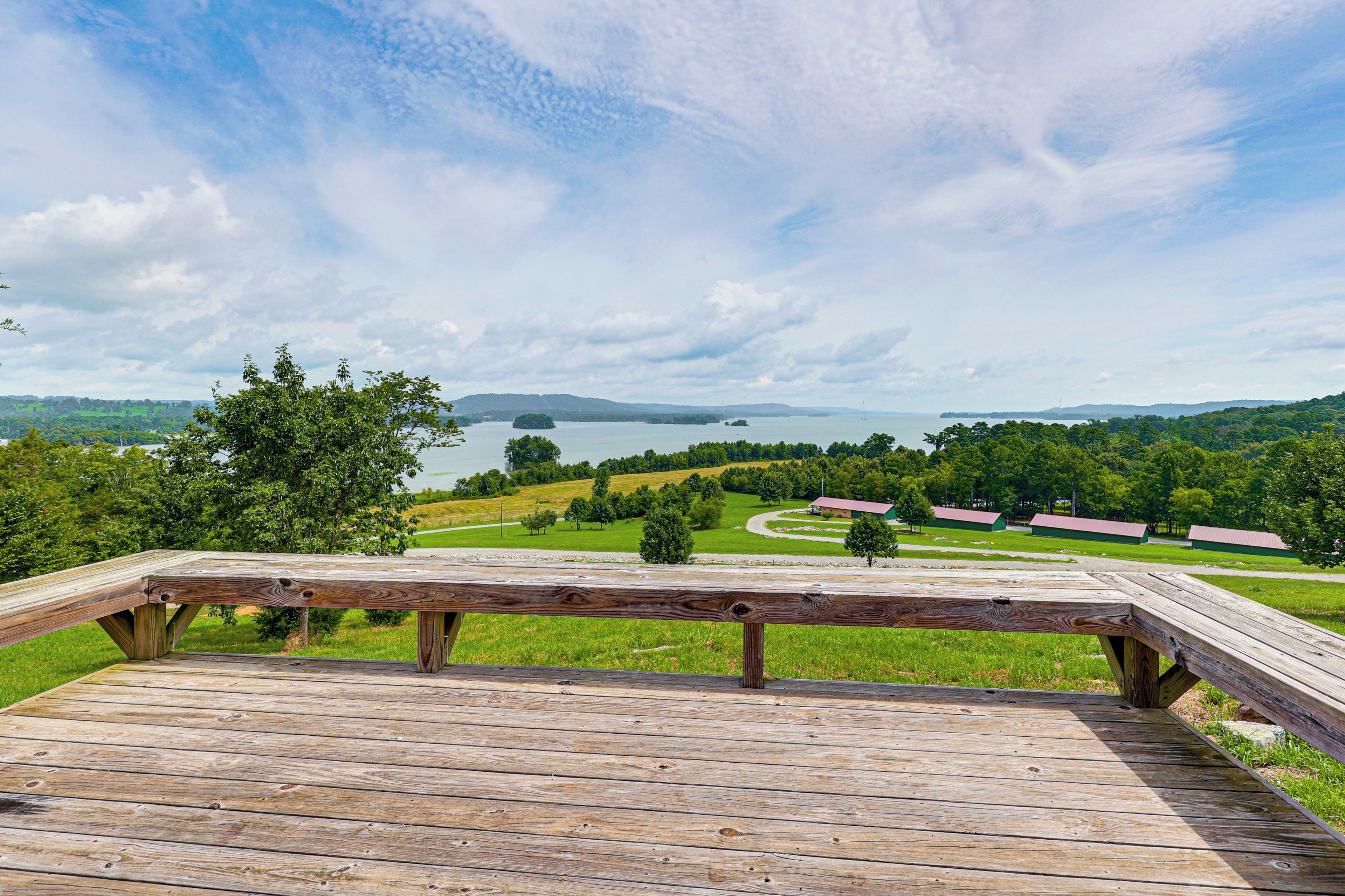 A wooden deck with a view of a lake, green fields, and distant buildings.
