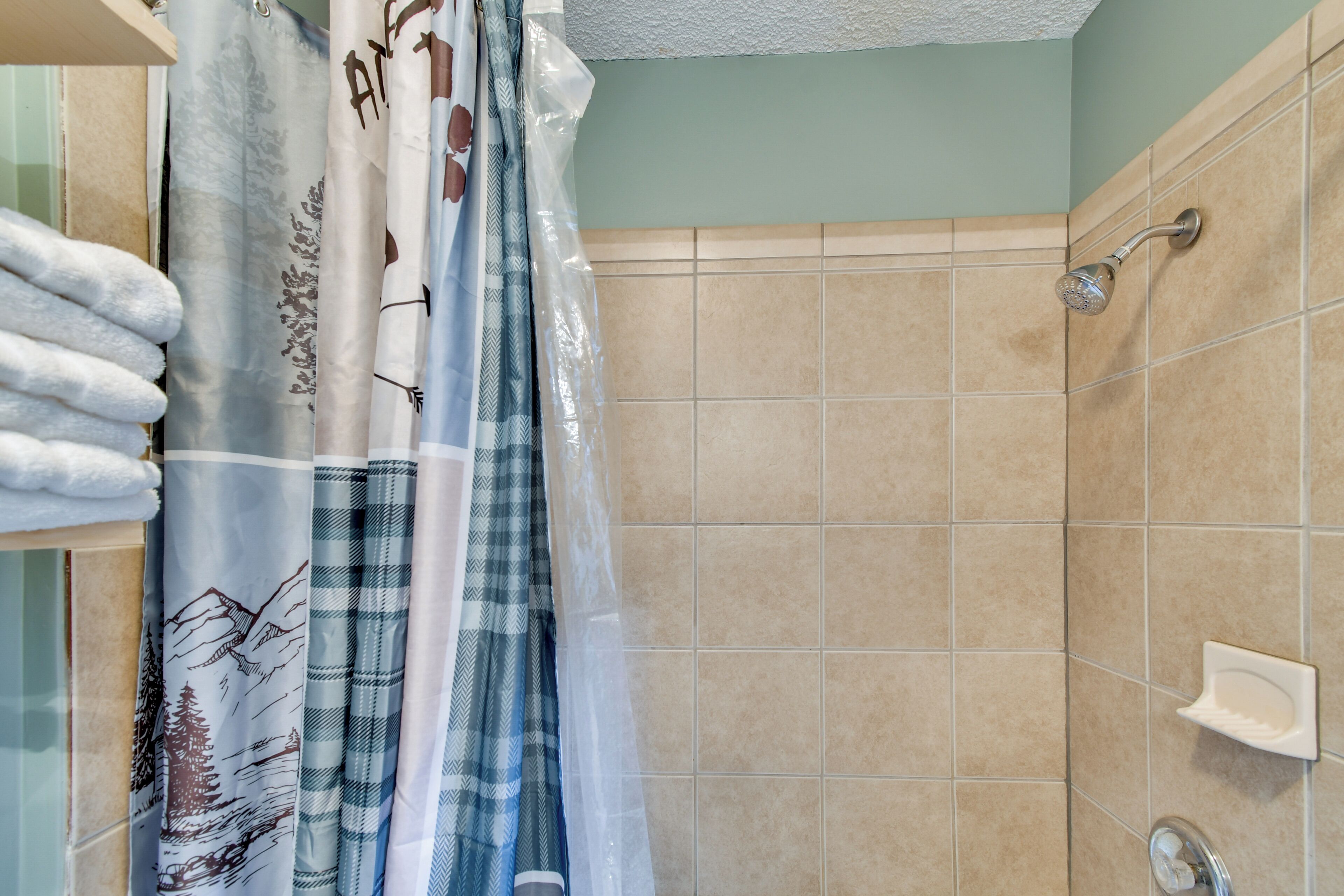 Un baño con baldosas color beige, una cortina de ducha con diseño de montaña y un jabonera blanca en la pared.