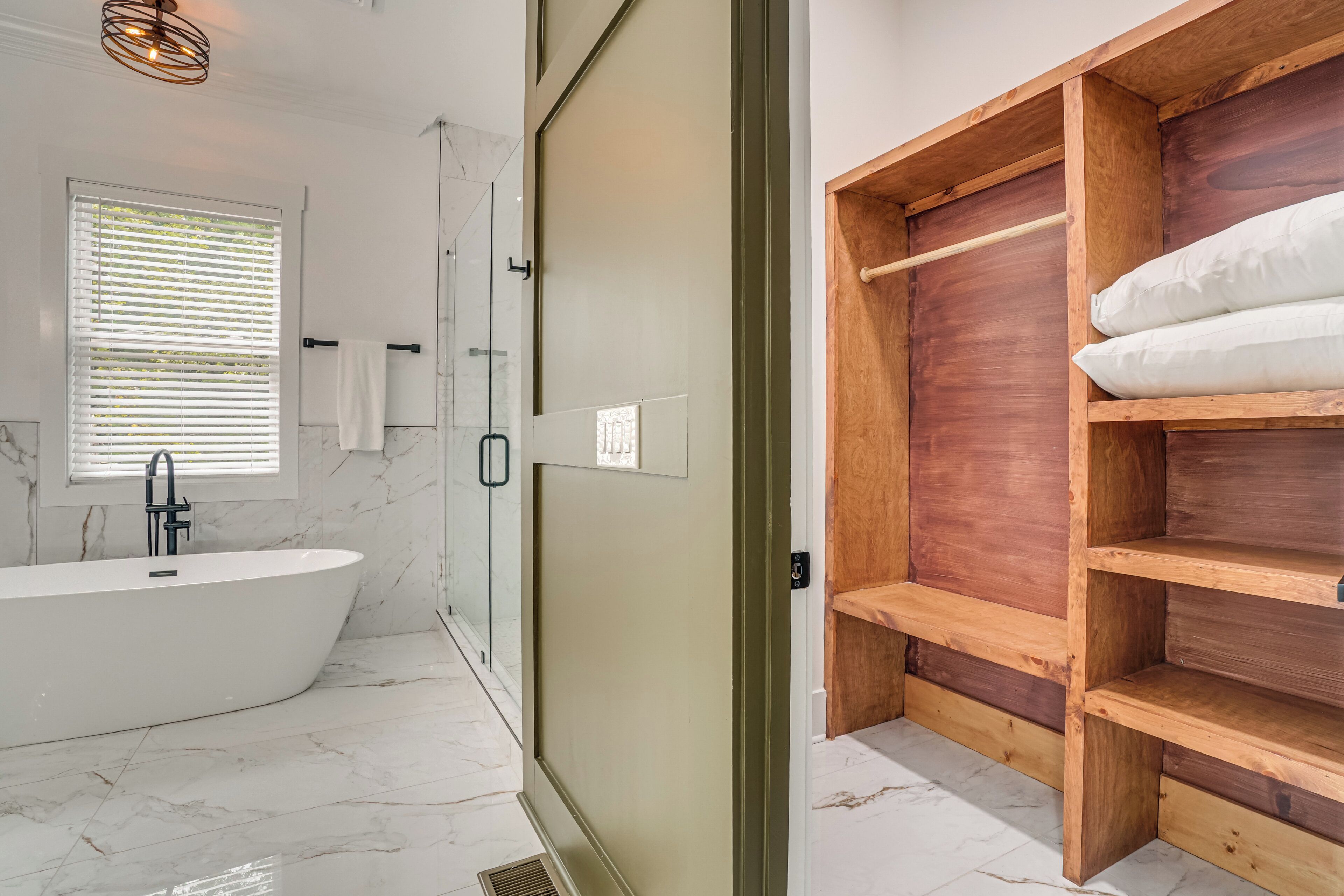 A modern bathroom with a freestanding bathtub, a glass shower enclosure, and a wooden shelf unit with a white pillow.