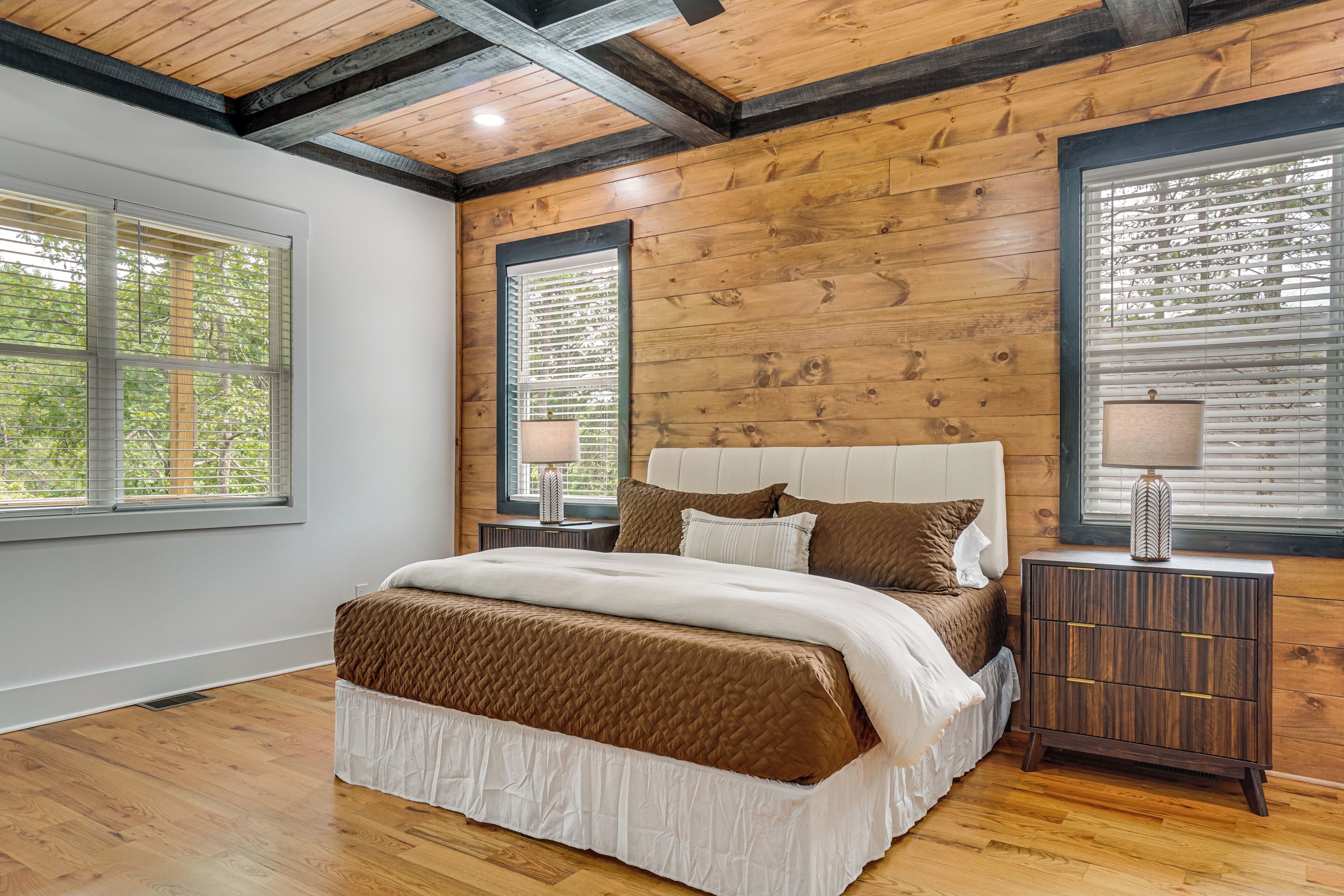 Une chambre avec un plafond en bois, un grand lit doté d’une tête de lit matelassée, deux tables de chevet avec des lampes et de grandes fenêtres équipées de stores.