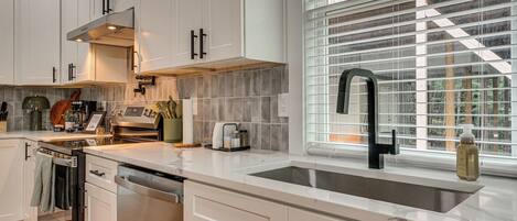 A modern kitchen with white cabinetry, a stainless steel dishwasher, a built-in oven, a black faucet, and a window with blinds.