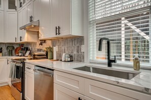Eine moderne Küche mit weißen Schränken, einem Edelstahl-Geschirrspüler, einem eingebauten Backofen, einem schwarzen Wasserhahn und einem Fenster mit Jalousien.
