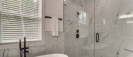 A modern bathroom with a freestanding bathtub, a glass-enclosed shower, and a window with blinds.