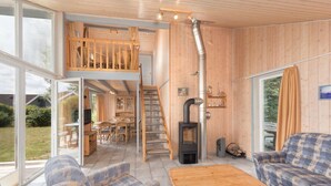 A spacious living area with a high ceiling, wooden walls, a loft bedroom, a fireplace, and a dining area. - Gemutliches Ferienhaus mit Garten und Terrasse im Hafendorf Muritz (Rechlin)