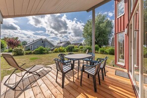 Eine Holzterrasse mit rundem Tisch und Stühlen, einem Liegestuhl und Blick auf Häuser im Hintergrund.