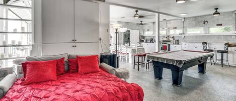 A room with a pool table, a bed with red bedding, and a kitchen area.
