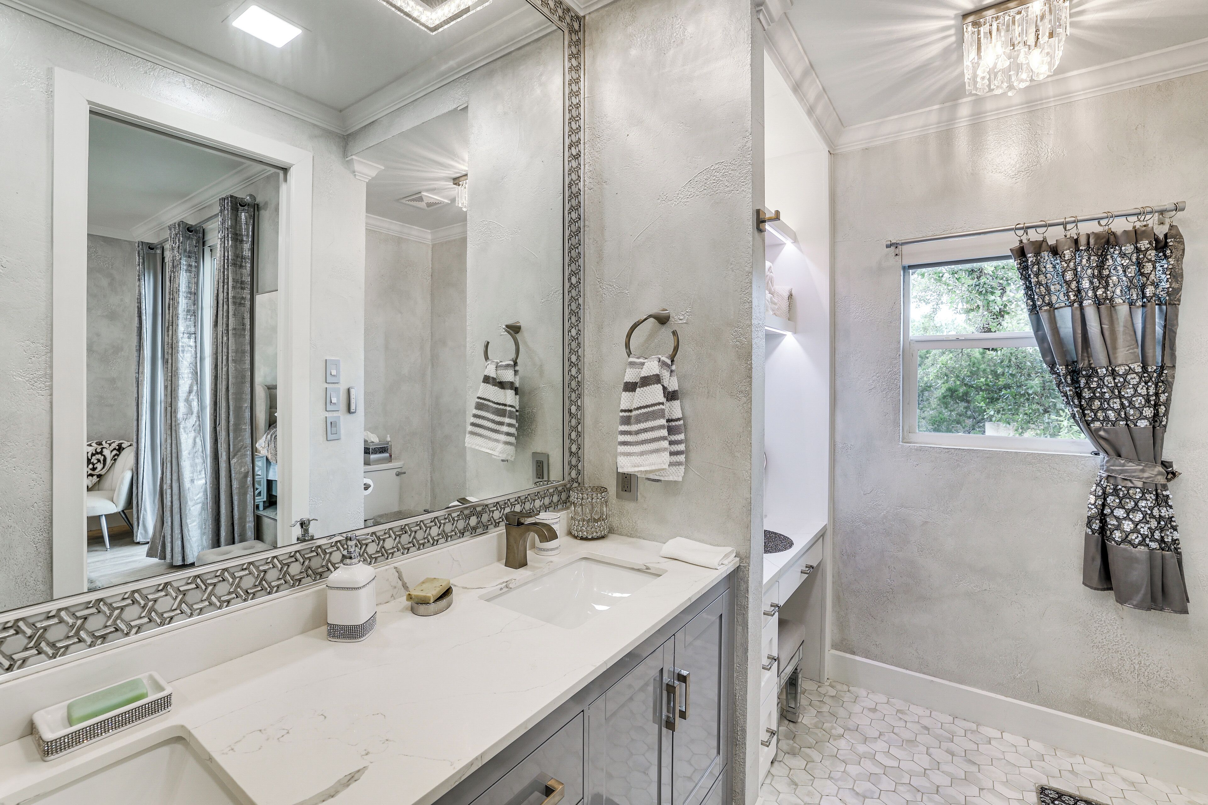 Une salle de bain dotée d’un grand miroir, d’un lavabo avec un plan de travail en marbre, d’un porte-serviettes et d’une fenêtre avec des rideaux.