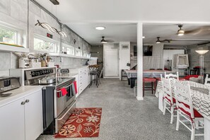 A modern kitchen with stainless steel appliances, a dining area with white chairs and a patterned rug, and a visible door leading to another room.