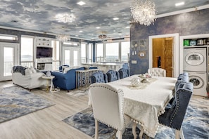 A spacious room with a large dining table, seating, a kitchen area, and a view through large windows.