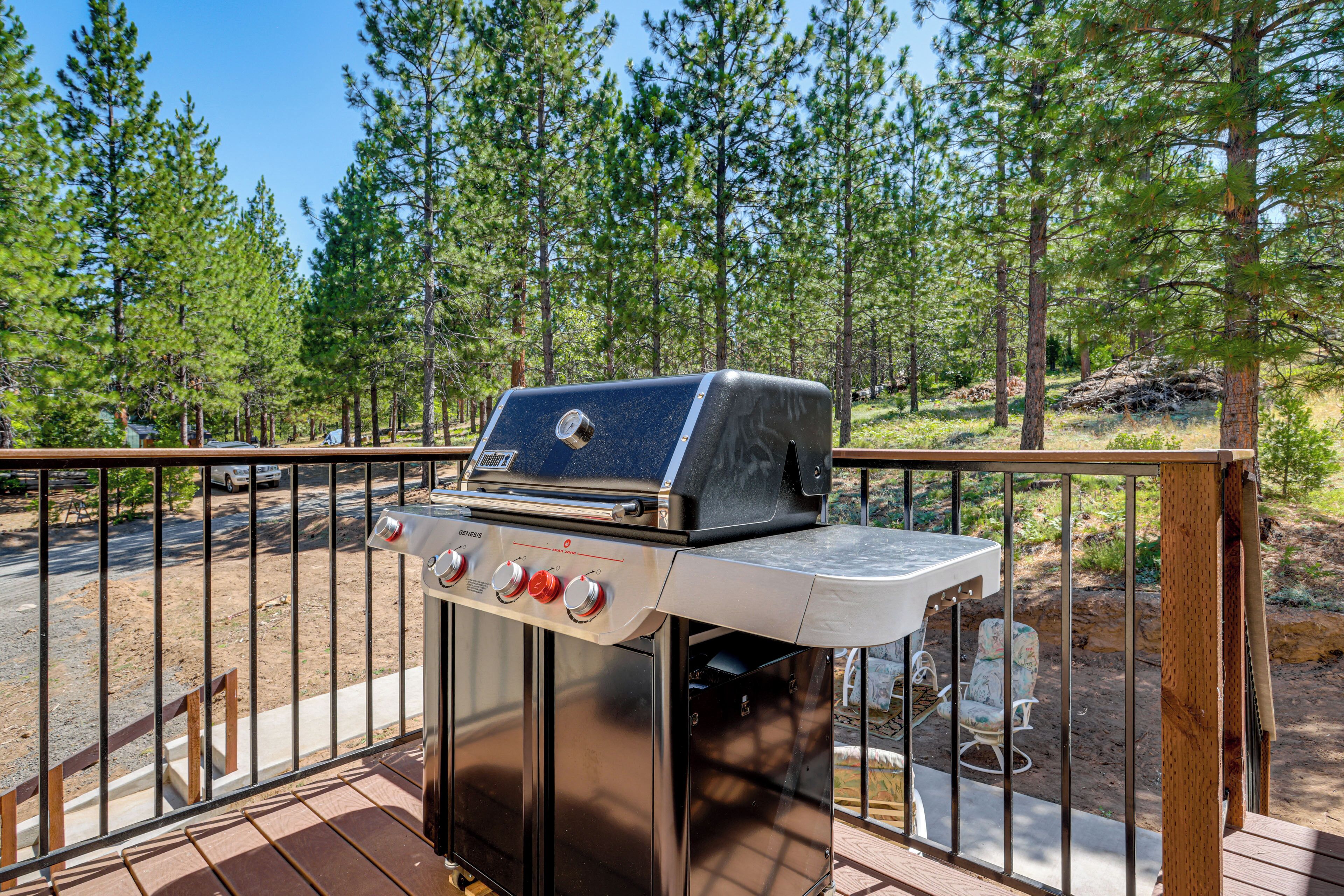 A barbecue grill on a wooden deck with a railing, surrounded by a forested area.