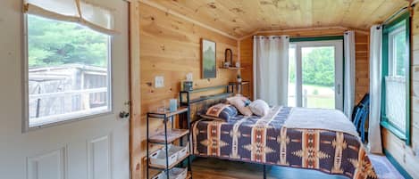 A wooden cabin bedroom with a bed, shelves, and a view of the outdoors.