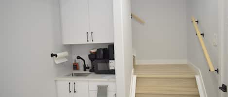 A small kitchen with white cabinets, a microwave, a coffee maker, and a wooden staircase.