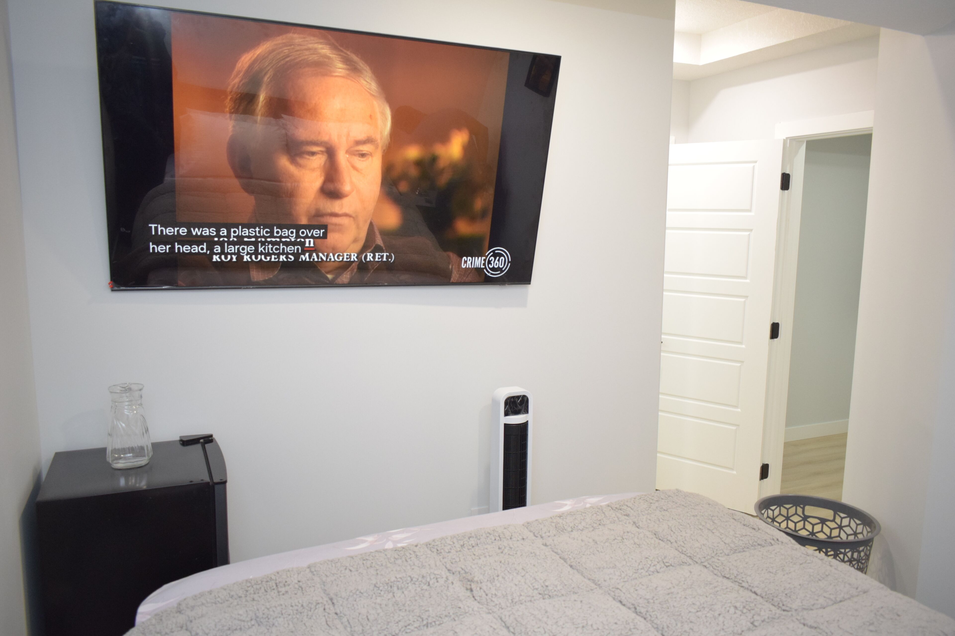 Quarto com cama, televisão na parede, mesa de cabeceira com um copo e porta para outro cômodo.