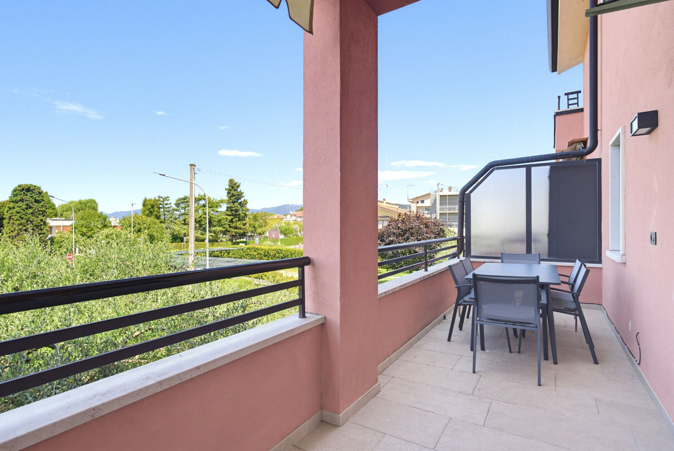 Uma varanda com mesa e cadeiras, vista para vegetação e edifícios, e fachada rosa de um prédio.