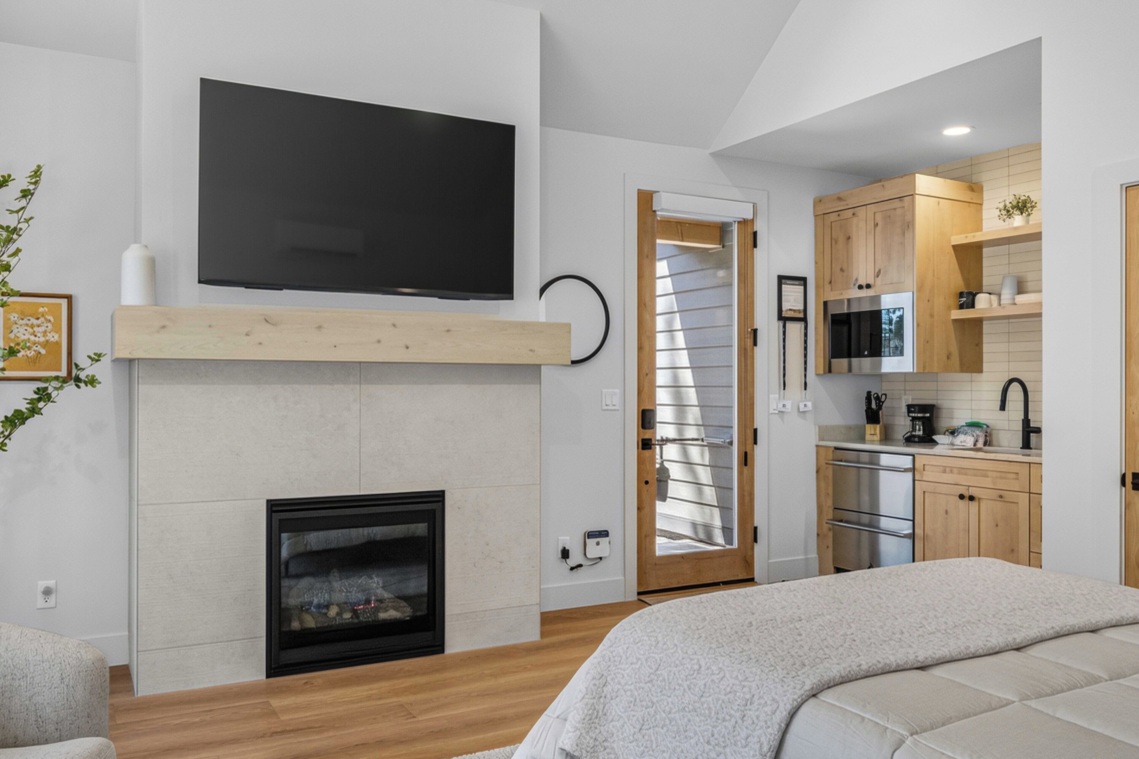 A modern interior with a fireplace, a flat-screen TV mounted above it, a kitchen area with wooden cabinets, and a bed with a white blanket.