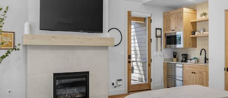 A modern interior with a fireplace, a flat-screen TV mounted above it, a kitchen area with wooden cabinets, and a bed with a white blanket.