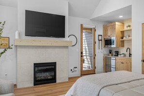 Un interior moderno con chimenea, un televisor de pantalla plana montado encima, una zona de cocina con armarios de madera y una cama con manta blanca.