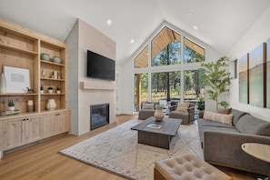 A modern living room with a fireplace, large windows, and a television mounted on the wall. - NL33A | 56457-1 Elk Run Drive (Sunriver)