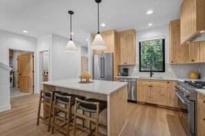 A modern kitchen with wooden cabinets, a stainless steel refrigerator, a built-in oven, and a large window offering a view of trees. - NL33A | 56457-1 Elk Run Drive (Sunriver)