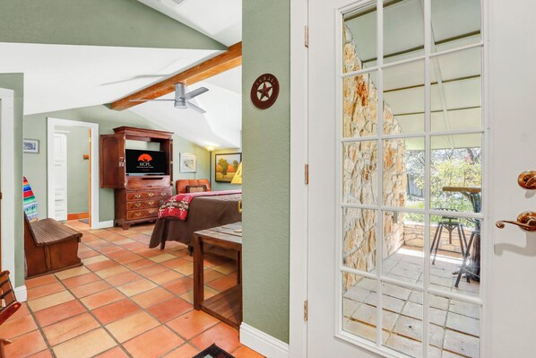 A room with a wooden cabinet, a television, a bed, a dining table, and a large window with a stone wall. - Hillsedge Haciendas | Multi unit Hill Country Retreat with Hot Tubs (Wimberley)