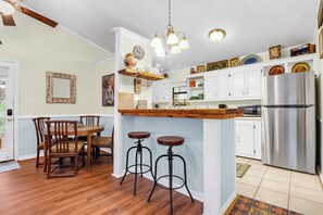 A kitchen with white cabinets, a stainless steel refrigerator, wooden stools, and a hanging light fixture. - Hillsedge Haciendas | Multi unit Hill Country Retreat with Hot Tubs (Wimberley)