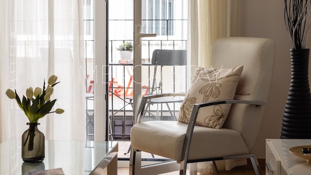 Ein modernes Wohnzimmer mit einer Couch, einem Glastisch, einer Vase mit Blumen und Blick auf den Außenbalkon.