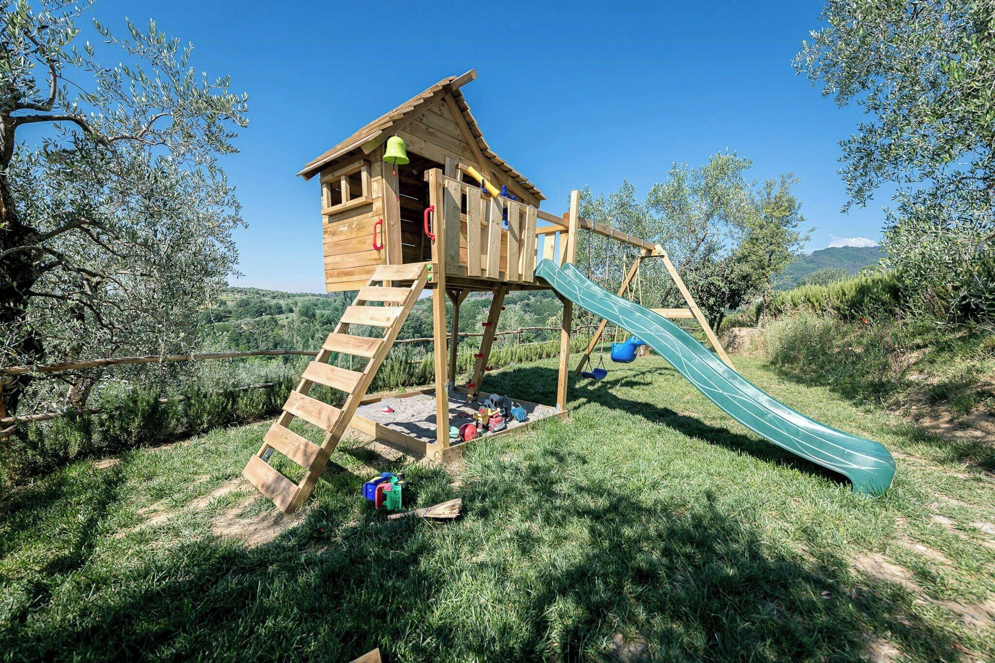 Une cabane en bois avec un toboggan et une zone d’escalade, entourée d’herbe et d’arbres.