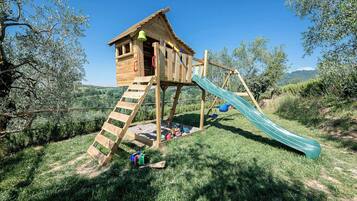 Une cabane en bois avec un toboggan et une zone d’escalade, entourée d’herbe et d’arbres.
