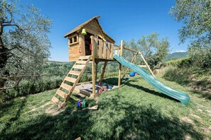 Una casita de juegos de madera con un tobogán y una zona de escalada, rodeada de césped y árboles.