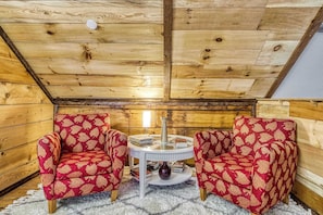 Ein gemütlicher Raum mit Holzwänden und schrägverdeckelter Decke, mit zwei rot gemusterten Sesseln und einem kleinen runden Tisch mit Büchern und einer Vase.