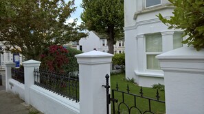 Ein weißes Gebäude mit schwarzem Eisenzaun, ein Baum mit roten Blüten und einer gepflegten Rasenfläche.