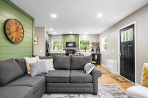 Ein modernes Wohnzimmer mit Ecksofa, einer großen Wanduhr und einer Küche im Hintergrund.