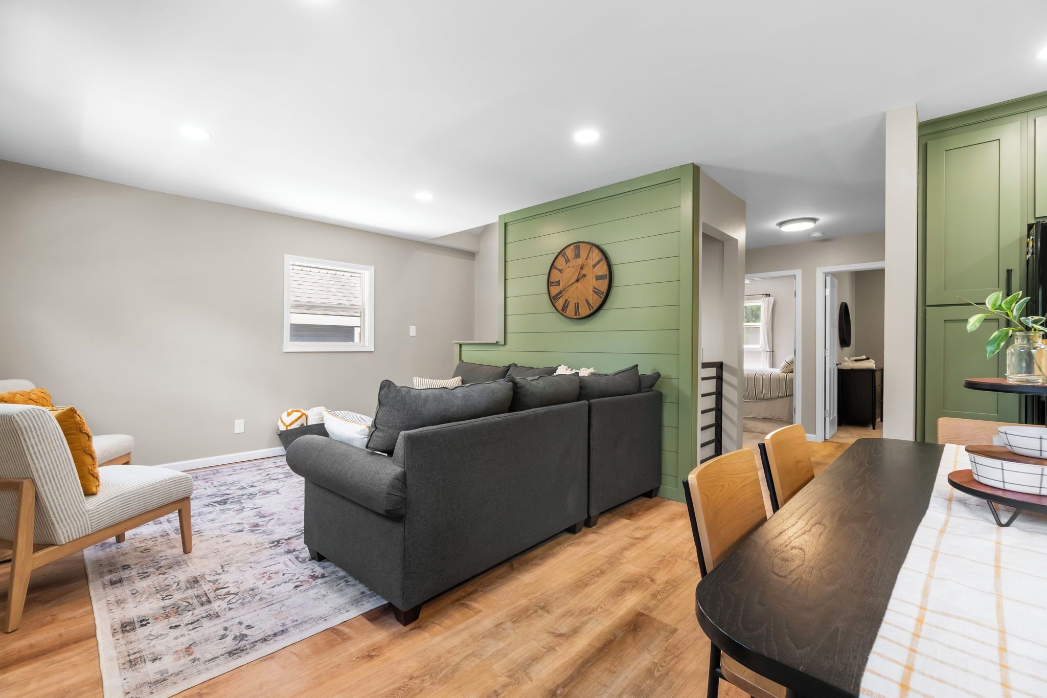 A modern living room with a grey sofa, a dining table with chairs, a wooden floor, and a green accent wall.