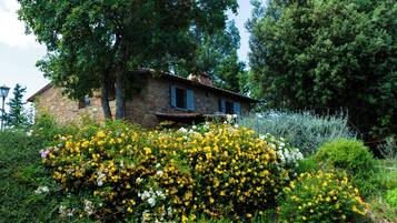 Una casa in pietra con tetto spiovente, circondata da vegetazione lussureggiante e cespugli fioriti.