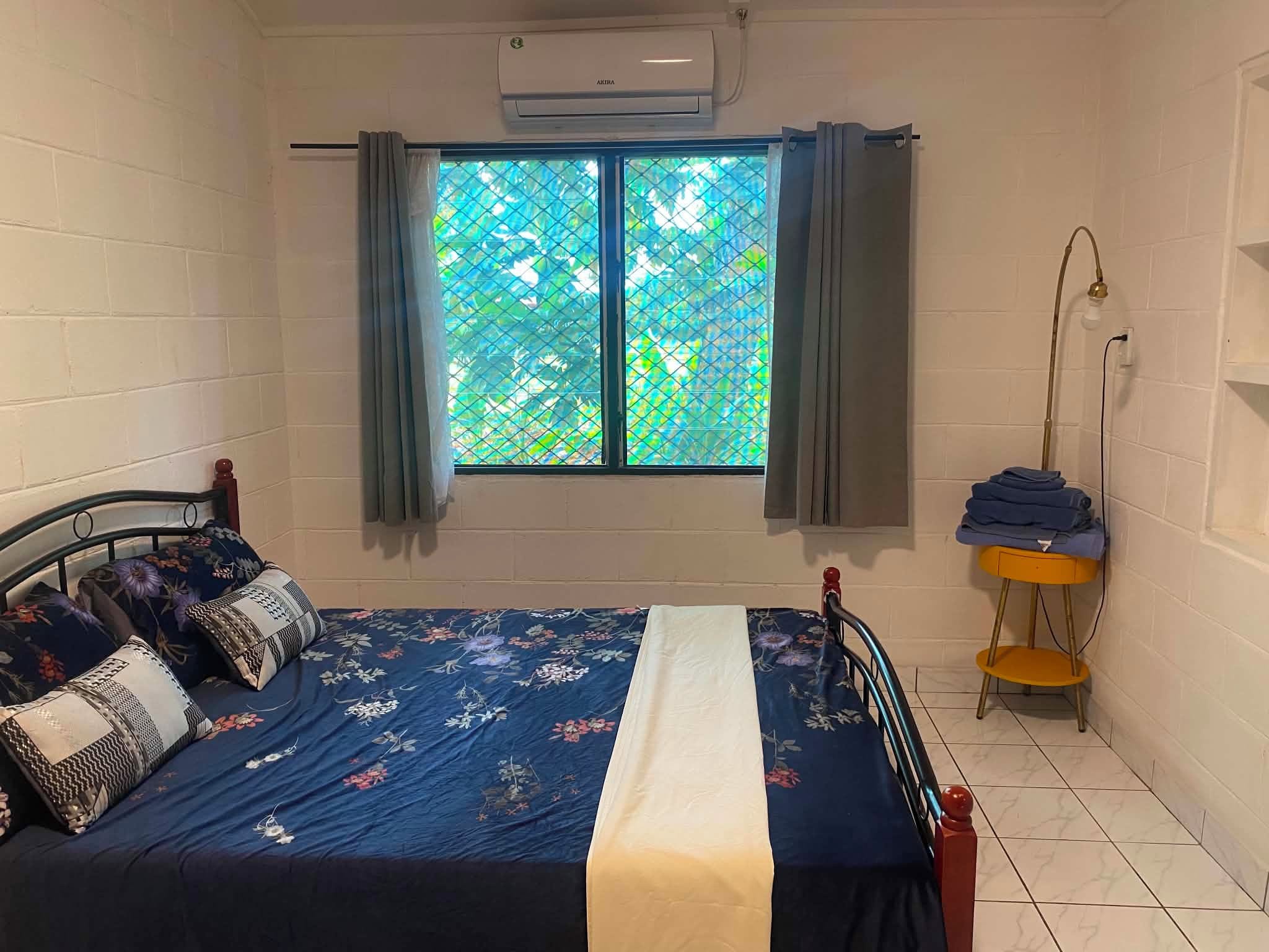 Ein Schlafzimmer mit Bett, einem Fenster mit Vorhängen, einer Stehlampe und einem kleinen Tisch mit gefalteter Kleidung.