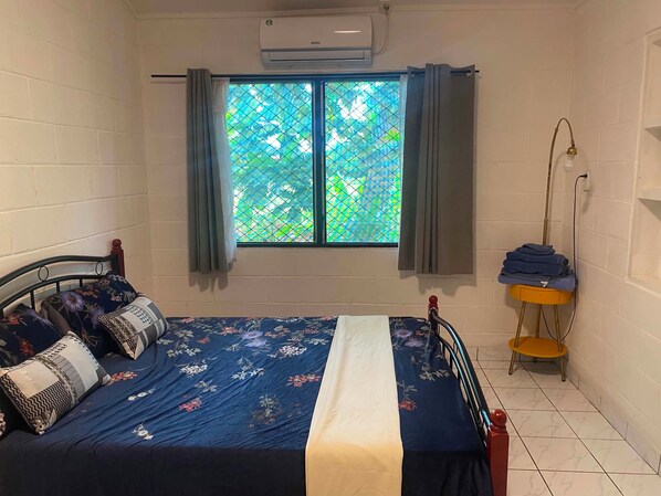 A bedroom with a bed, a window with curtains, a standing lamp, and a small table with folded clothes.