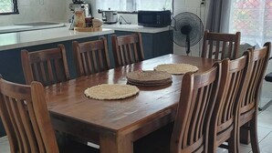A dining area with a wooden table and chairs, a kitchen in the background, and a window with curtains. - Home in Siusega - Vonnys Garden House (Vaitele)