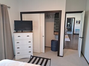 Ein Schlafzimmer mit Fernseher auf einer Kommode, einem Bett, einem Sessel, einem Kleiderschrank und einer Tür zu einem anderen Raum.