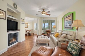 A spacious living room with a fireplace, leather furniture, a ceiling fan, and large windows. - Havens 1136 Back on Program! Beautiful & Spacious! In Barefoot Resort! (North Myrtle Beach)