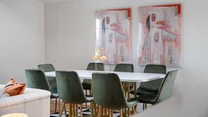 Un comedor con una mesa blanca y sillas verdes, dos cuadros abstractos en la pared y una mesita con una lámpara.