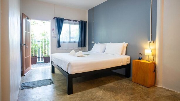 A bedroom with a bed, a bedside table with a lamp, a window with curtains, and a door leading to an outdoor area. - Fix25House Koh Kood (Ko Kood)