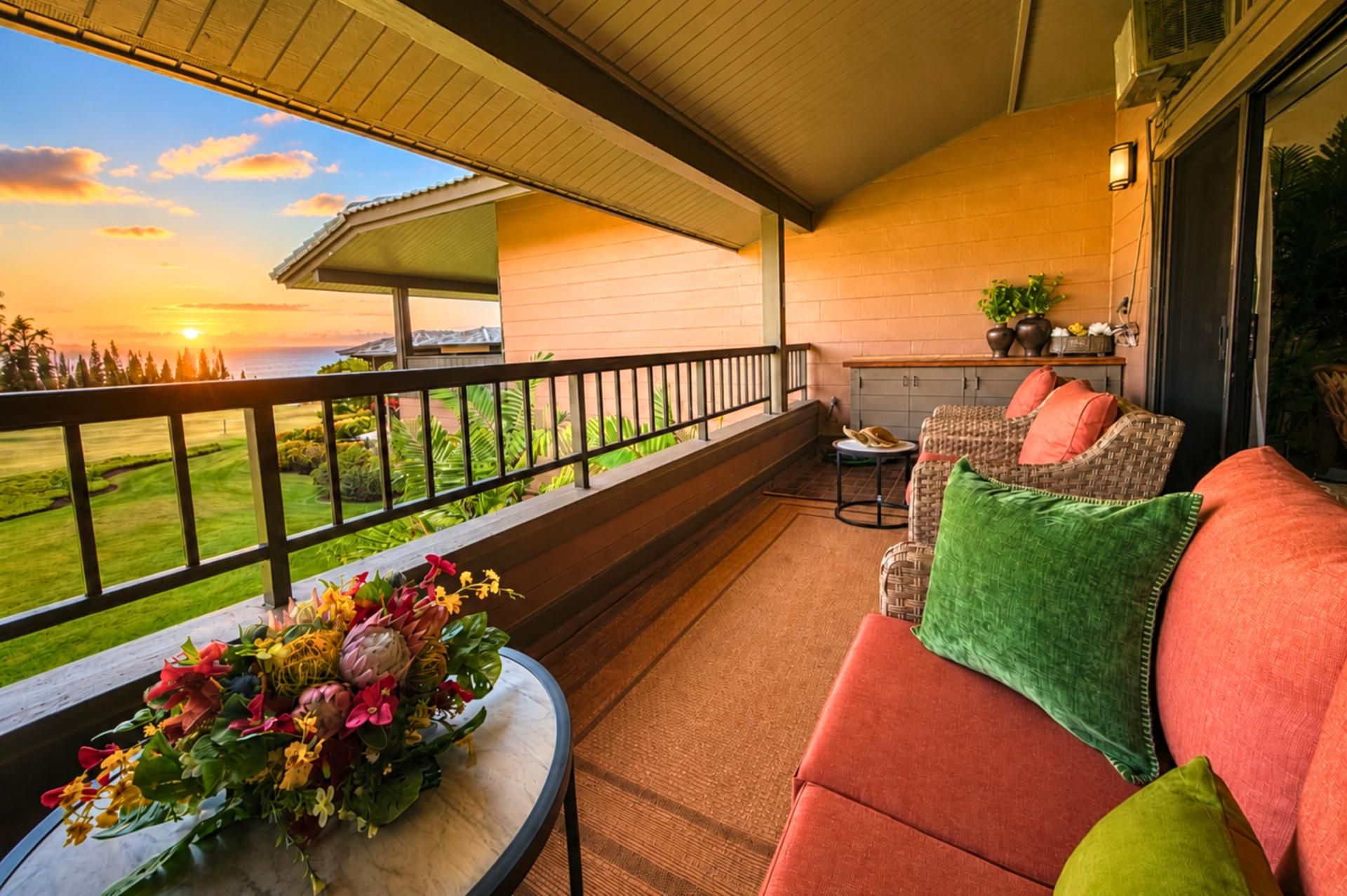 A balcony with a view of the sunset, a wicker sofa with cushions, and a table with a floral arrangement.